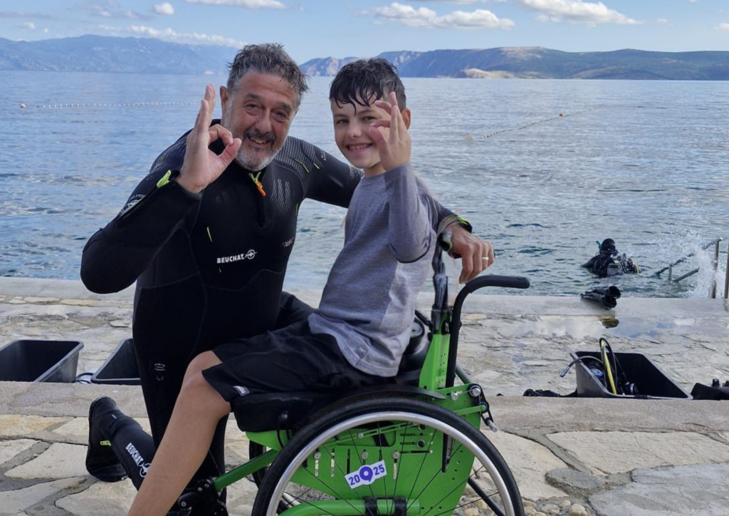 Tauchen mit Handicap - Ein Junge im Rollstuhl am Tauchen und der Tauchlehrer kniet daneben 